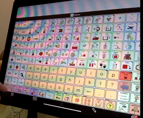 Figure 2 is a picture of an iPad with the program Proloquo2Go. The program shows a grid of buttons with varying symbols denoting words. On the bottom of the grid there is a QWERTY keyboard laid out in yellow buttons (to visually pop compared to the usually-white button backgrounds).