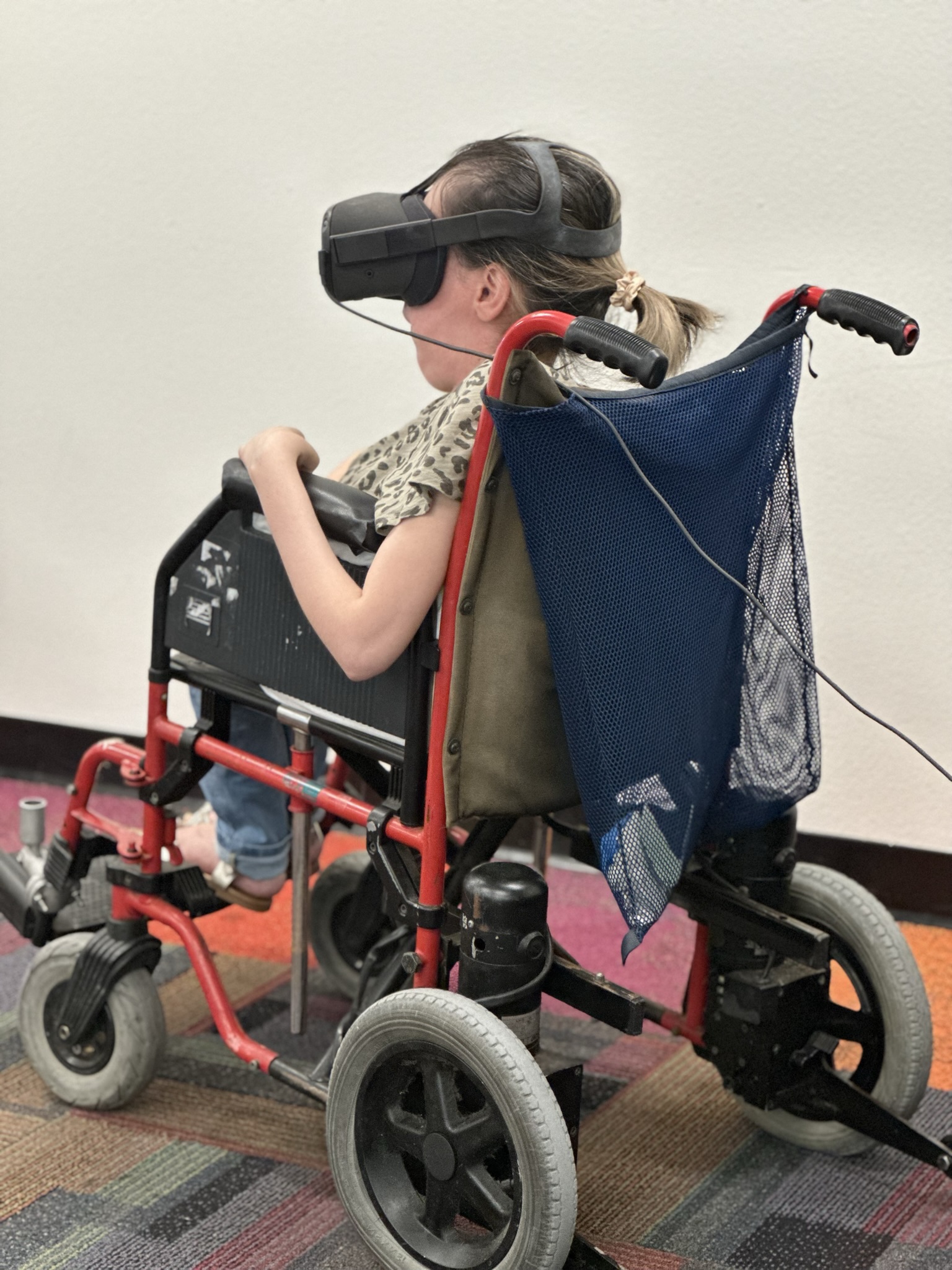 A person seated in a wheelchair, wearing a VR headset. The setting is an indoor space with multicolored carpeted flooring.
