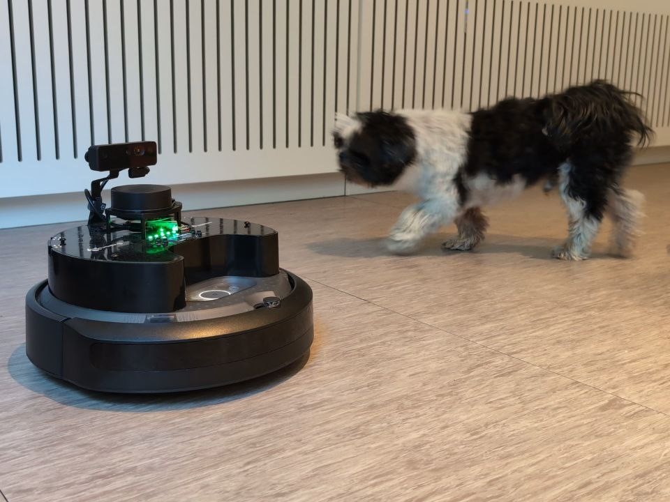 The TurtleBot robot, which looks like a vacuum cleaner robot, on the floor, and a dog next to it.