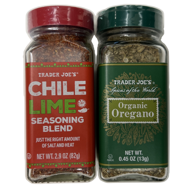 This image shows two clear glass spice jars from Trader Joe’s placed side by side. Each jar has a plastic screw-on lid and a colorful label on the front. The jar on the left has a red lid and a bright red label with white and green text. It reads “Trader Joe’s Chile Lime Seasoning Blend” in bold letters, with the word “Chile” in white and “Lime” in a vivid green, which is accompanied by small icons of a lime slice and a chili pepper. Beneath that, it says “Just the right amount of salt and heat,” and it contains 2.9 ounces or 82 grams of spice mix. Inside the jar, the seasoning appears reddish-orange with visible flecks of chili and possibly dried lime zest. The jar on the right has a dark green lid and a matching green label with a decorative botanical pattern. It reads “Trader Joe’s Spices of the World Organic Oregano” in white and olive green text. The oregano inside is dry and pale green with small, crumbled leaves visible through the glass. This jar contains 0.45 ounces or 13 grams. Both jars are cleanly designed and stand upright, showing their contents clearly through the transparent glass.
